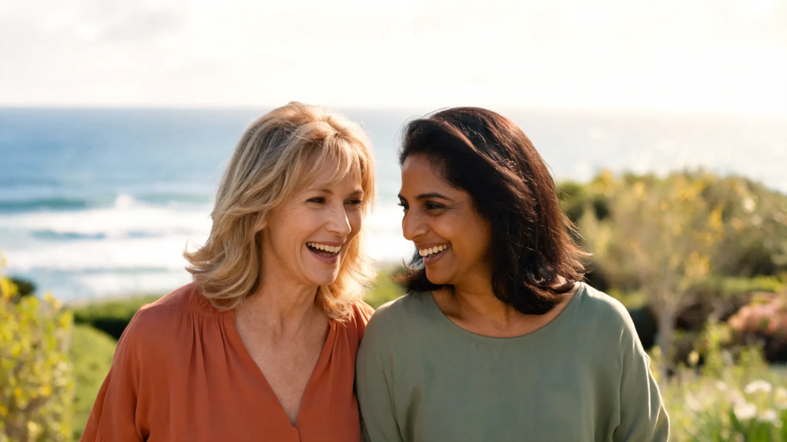 Two women in their sixties walking together and laughing on a coastal path in spring sunshine.
