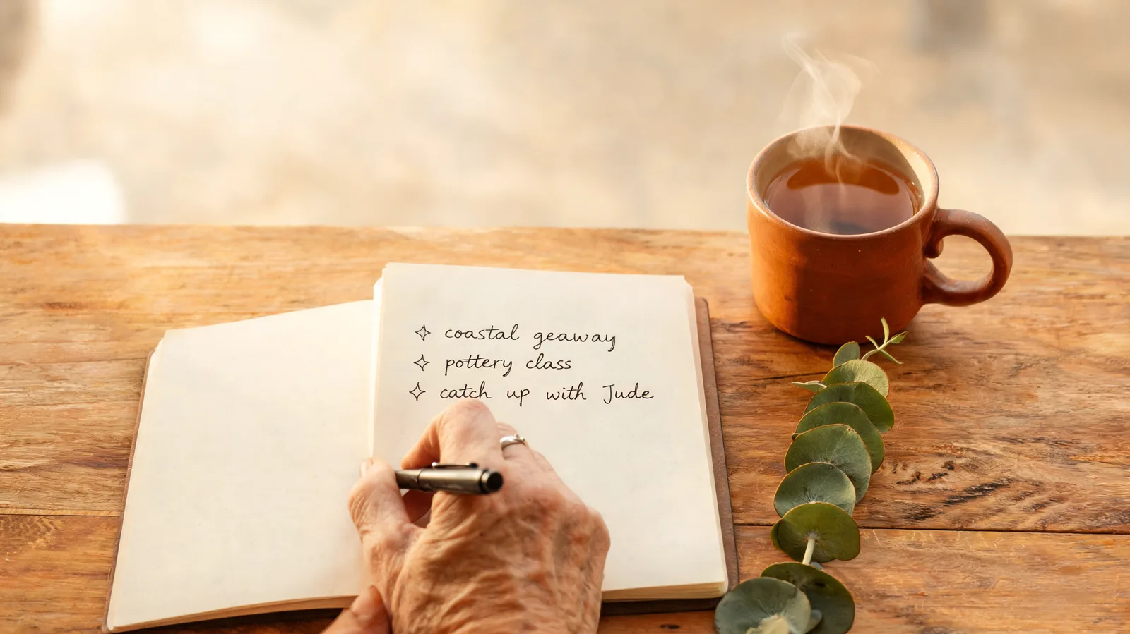 Older hands writing a short list in a notebook beside a cup of tea and a sprig of eucalyptus