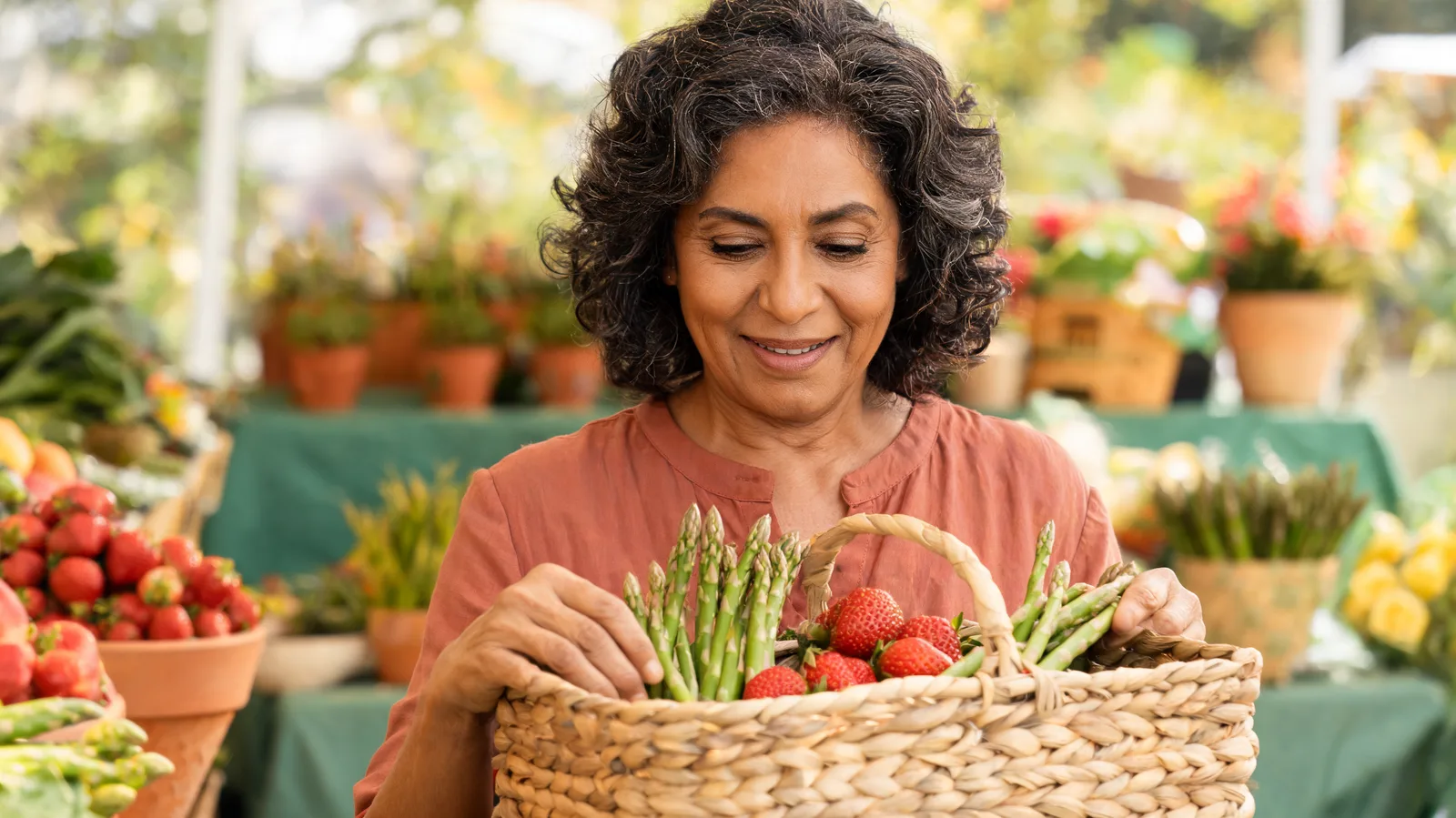 An older woman at a farmers market stall placing fresh asparagus and strawberries into a woven basket.