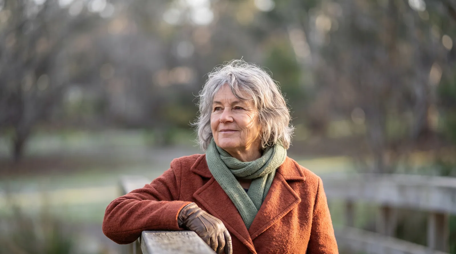 An older woman in a terracotta wool coat and sage green scarf resting a gloved hand on a timber handrail outdoors, with winter parkland softly blurred behind her.