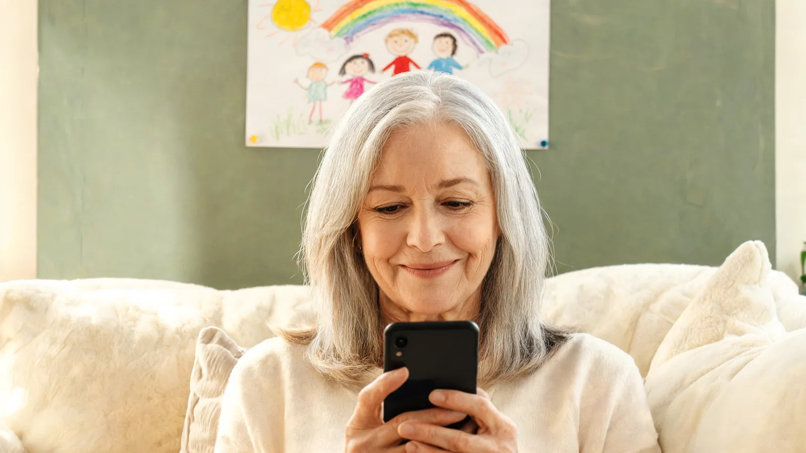 Older woman on her lounge smiling at her phone, a child's drawing on the wall behind her.